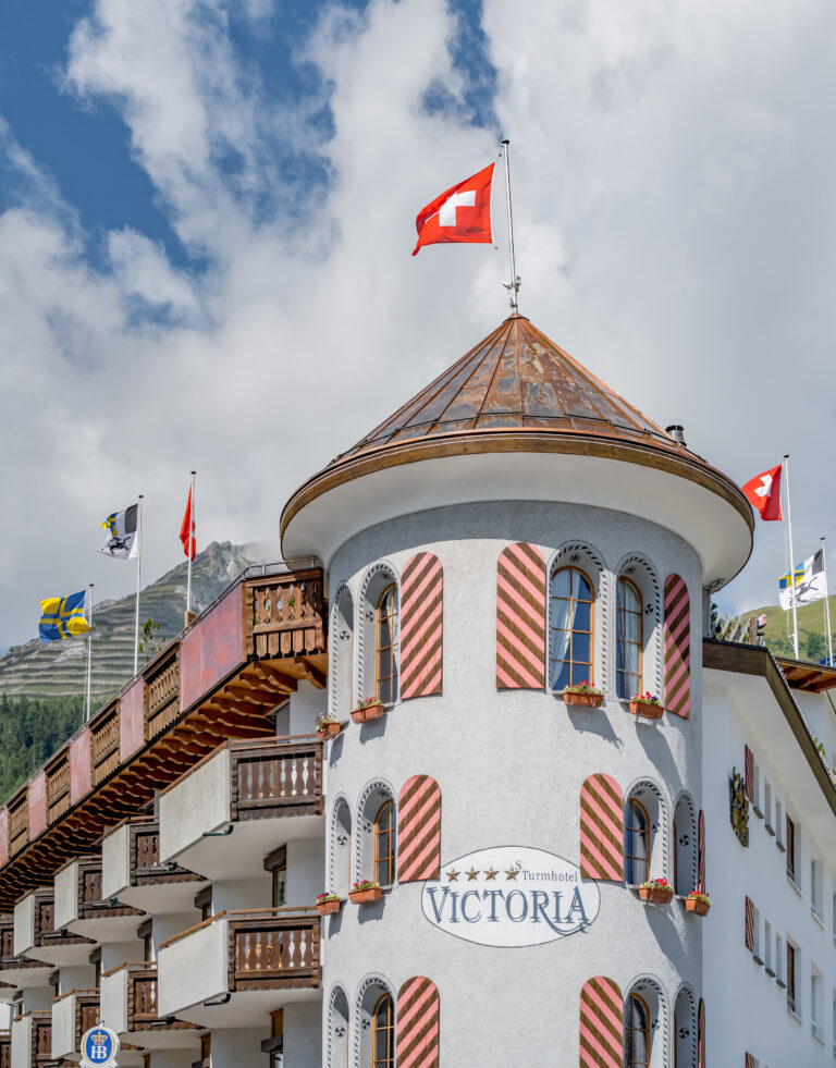 Turm des Turmhotels Victoria in Davos mit Schweizer Flagge vor blauem Himmel.